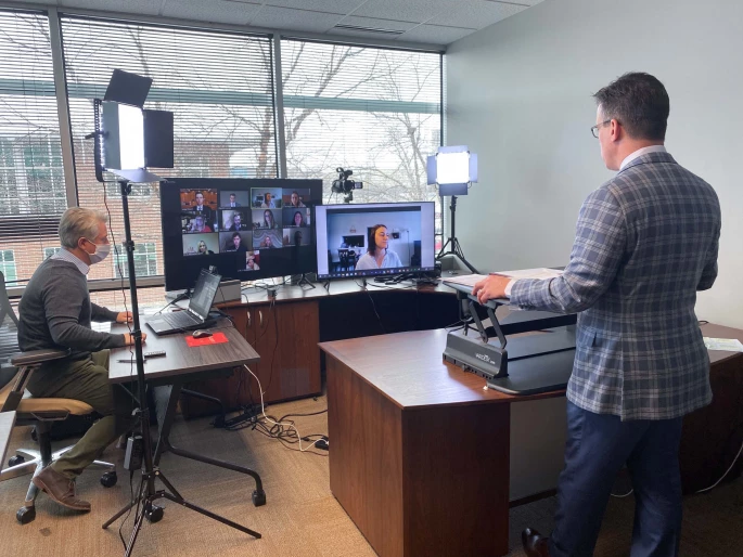 Two men in an office. on the left one sitting at a desk facing a laptop. on the right one standing in a suit facing monitors and lights in a virtual courtroom setting.