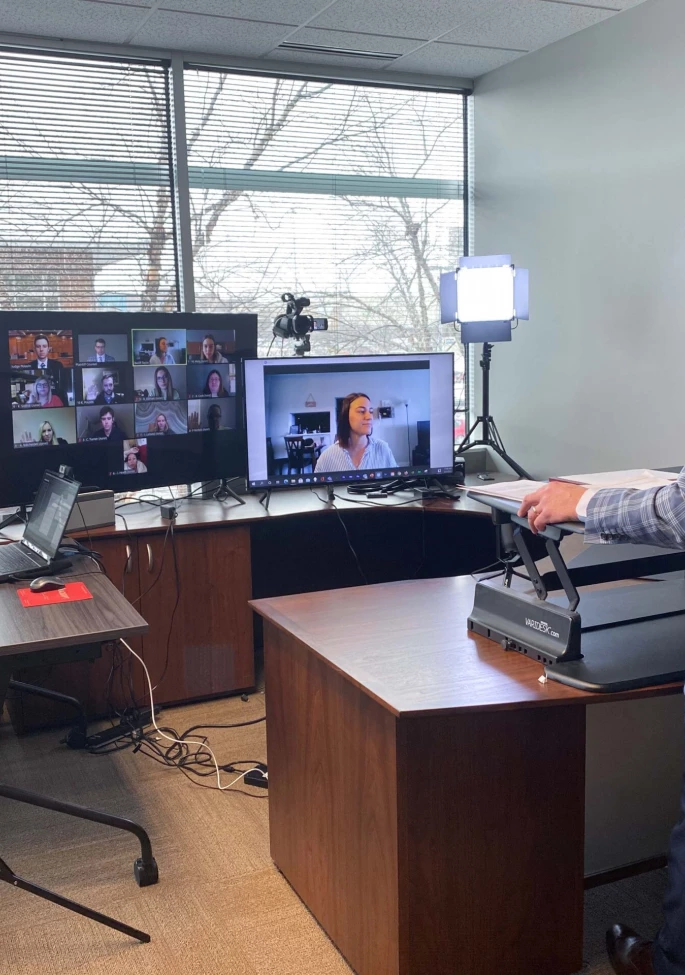 Two men in an office. on the left one sitting at a desk facing a laptop. on the right one standing in a suit facing monitors and lights in a virtual courtroom setting.